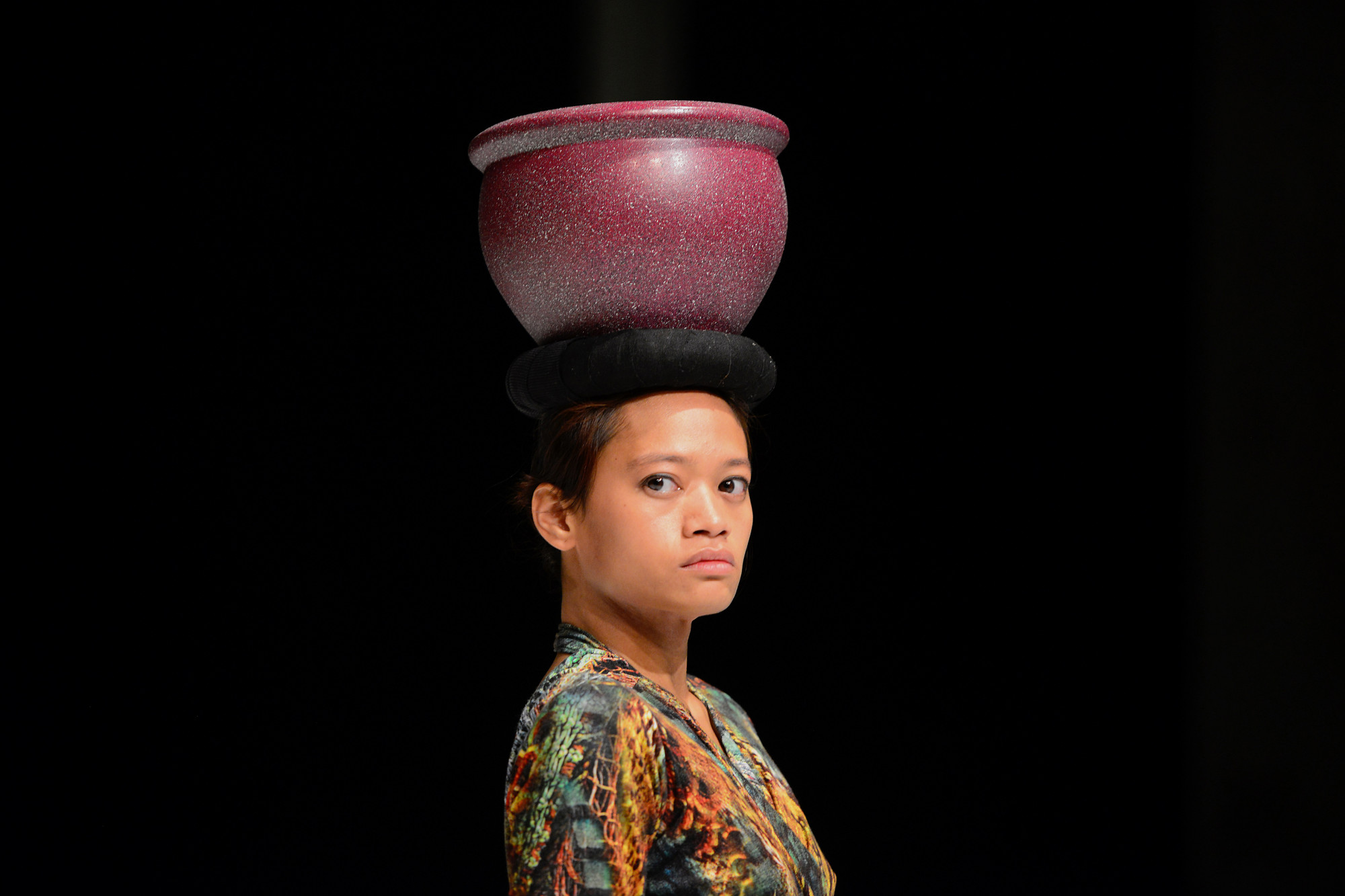 A young woman looks over her shoulder with a red vase balancing on her head.