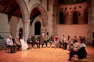 A group of people sitting in a semi-circle while 1 person is talking animatedly. They are in a dance studio with wooden floors in an old church.