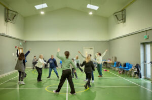 A group of women stand in a circle with one of their arms in the air. They are in a sports hall wiht a green floor. Everyone is moving.