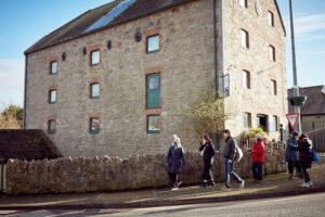 A group of people walking over a bridge with an old converted mill behind them. They are walking and talking, the photographer is across the road from them. The sky is blue.