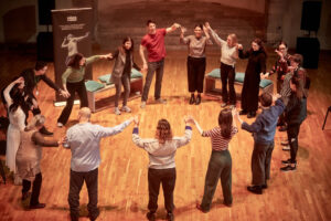 A group of people standing in a circle with their arms half raised and joined together by their fingers. The photo is taken from behind some of the group. They are in a dance studio with wooden floors in an old church.