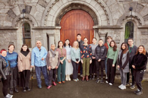 A group of 17 people standing outside, infront of the wooden doors of a church. There are a mix of men and women and everyone is smiling.