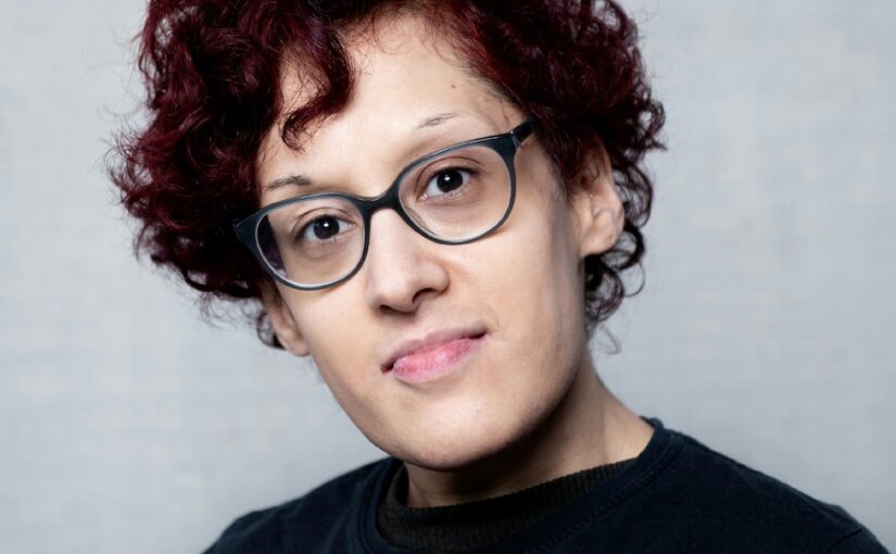 Headhot of a woman with short red curly hair and black glasses wearing a black jumper.