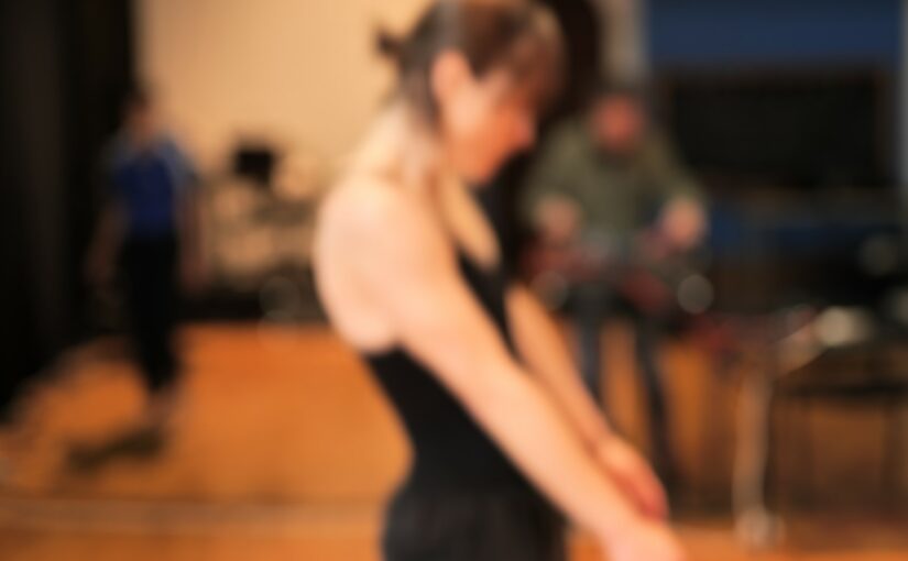 Blurry image of a dancer wearing a black t-shirt with her arms extended towards the front.