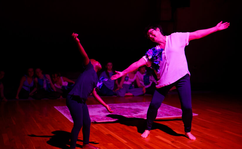 A young boy and a woman dancing together with their arms extended to their sides against a dark background.