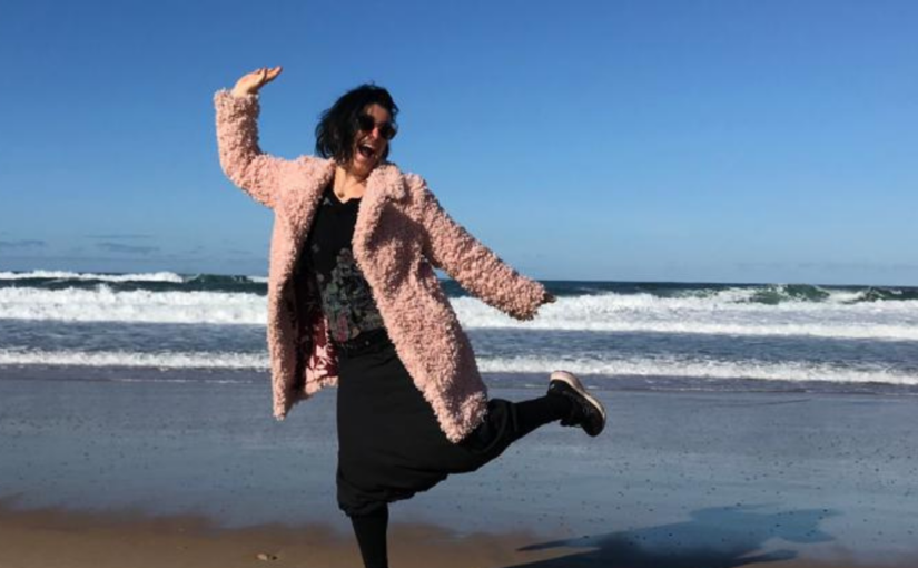 A woman wearing a black dress and a pink coat strikes a dancing pose on the beach against a blue sky.