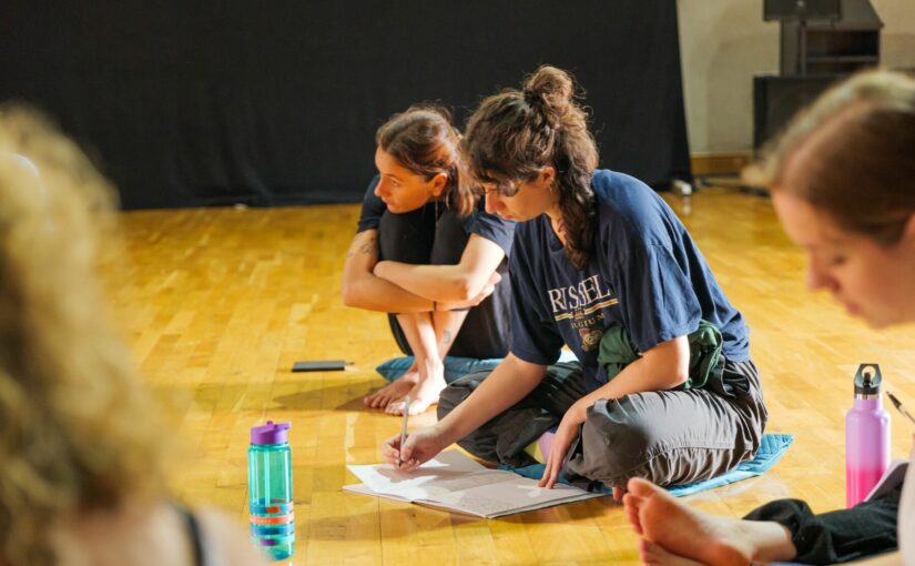 Three women sitting on the floor in comfortable clothes writing on a piece of paper while attending a class.