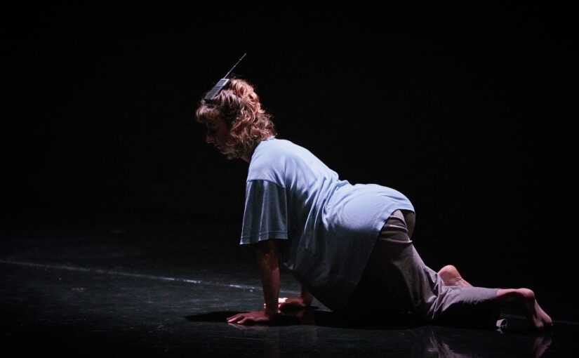 Image of a dancer on her hands and knees on the floor in a blue t-shirt.