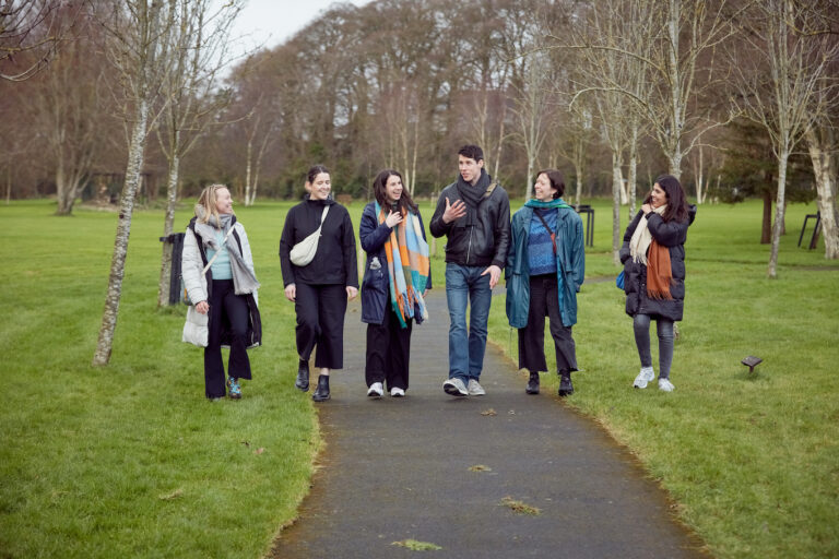 A group of 6 people walking in a line towards the camera. They are in a park, you can see the grey path beneath them, and the green grass beside them. Everyone is turned slightly towards the tallest person in the group who is speaking. The group are all wearing warm clothes as it is cold outside.