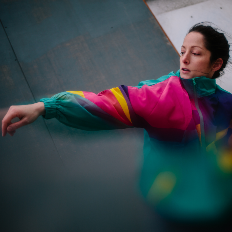 Image of a woman dancing in a bright coloured jacket, with one of her arms extended towards the front.