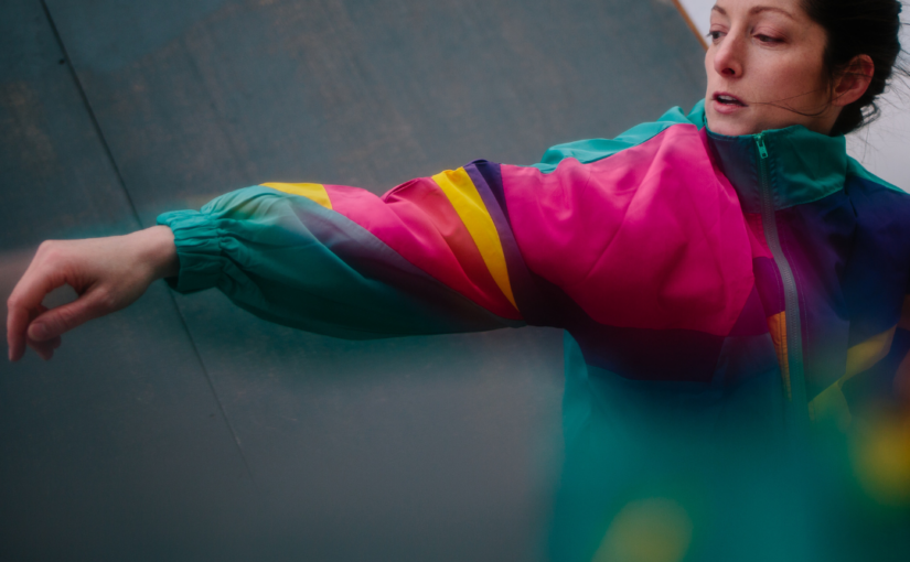 Image of a woman dancing in a bright coloured jacket, with one of her arms extended towards the front.