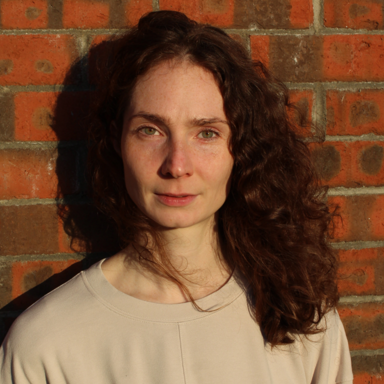 Headshot of a young woman with red curly hair agains a red brick background.