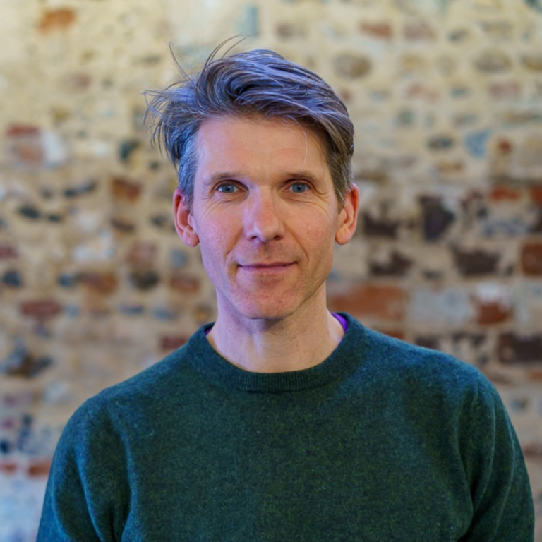 Headshot of a man wearing a dark green jumper against a red brick wall background.