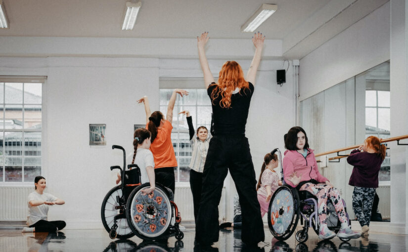 Image of a group of adults and children in a dance studio, some standing, some in wheelchairs, lifting their arms above their heads.