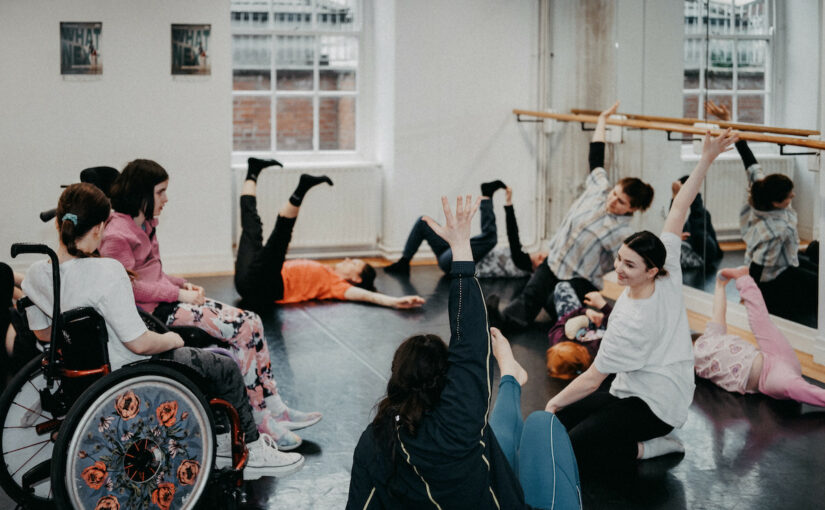 Image of a group of adults and children in a dance studio lying, sitting on the floor and in wheelchairs, lifting their hands and feet up.