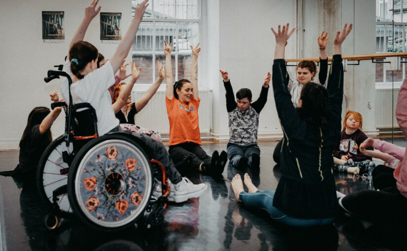 Image of a group of adults and children in a dance studio sitting on the floor and in wheelchairs, lifting their arms above their heads.