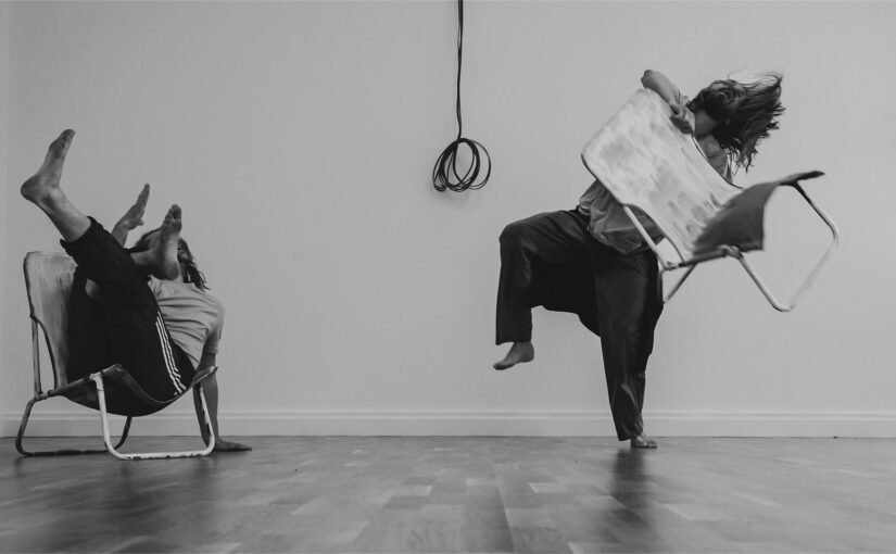 Black and white image showing a man holding a chair in the air and another one sitting on a chair.