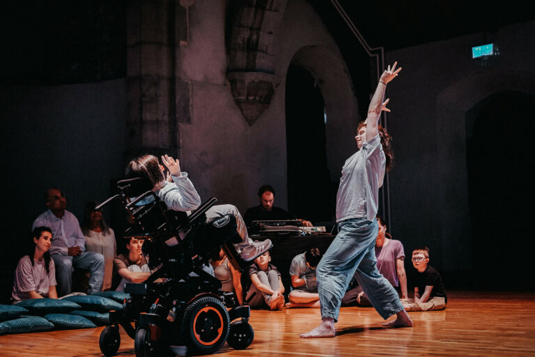 Image of two dancers doing a duet facing each other. One of them is using a wheelchair.