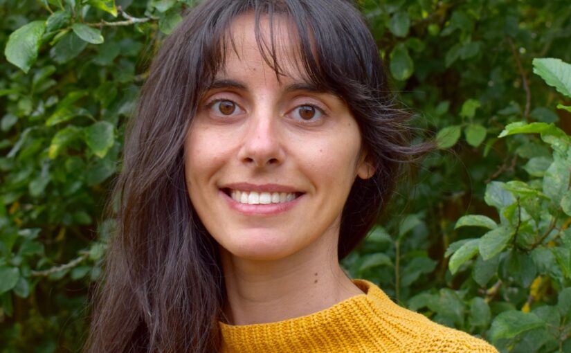 Headshot of a young woman with long brown hair wearing a yellow jumper against a background of green tree leaves.