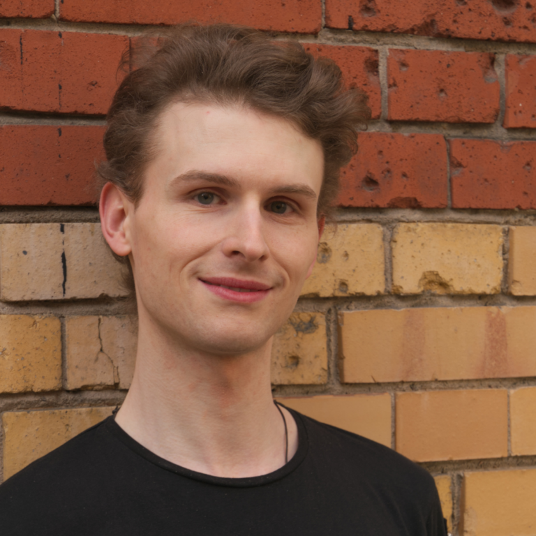 Headshot of a man with a black t-shirt against a yellow and red brick wall.