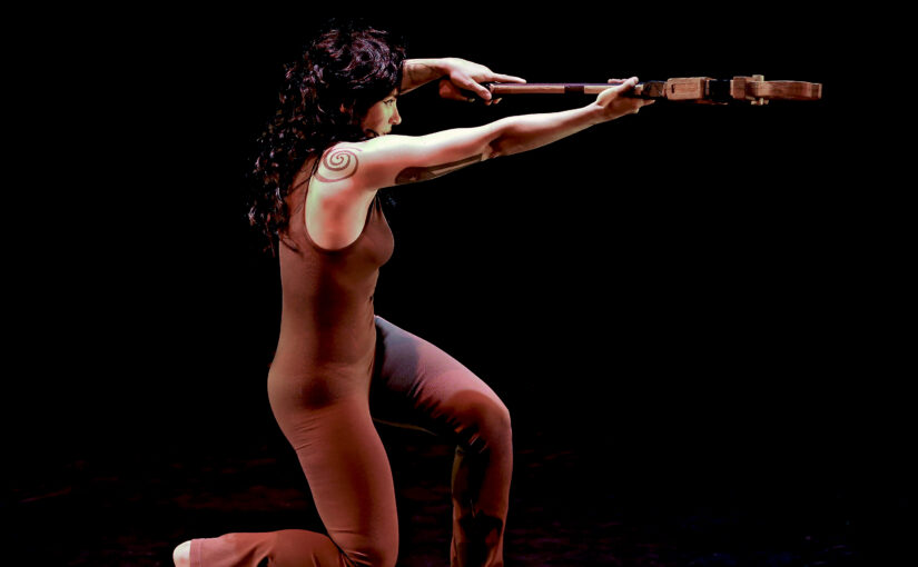 Image of a dancer with long dark hair and a brown bodysuit, holding a violin in front of them while kneeling.