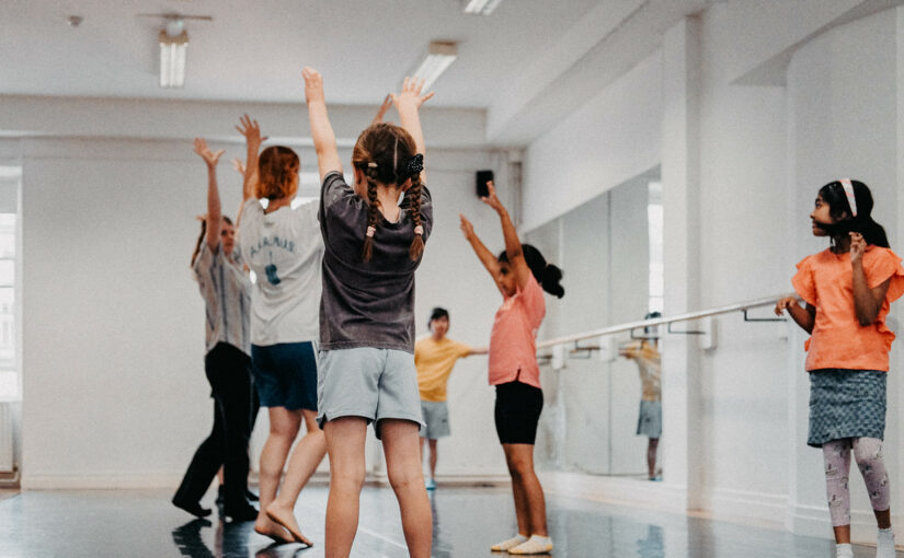 Image showing children and two teachers in a dance studio, all raising their hands up in the air.