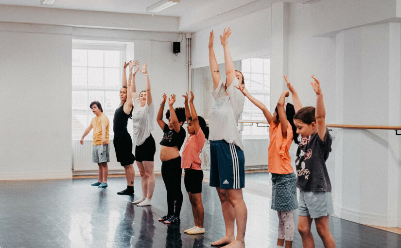 Image showing a group of children and two teachers during a dance class, all raising their arms up in the air.