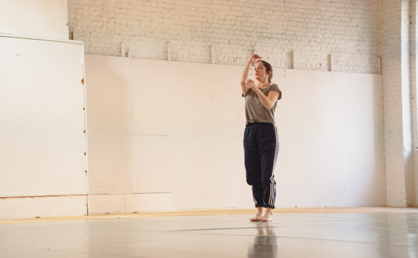 Image of a dancer standing with her hands in front of her, against a cream classroom background.