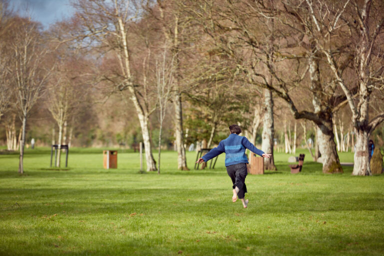 A person runs through a green park with lots of bare trees. We can the their back, they are weating a blue jumper and black jeans and have short brown hair. They are mid run and their legs don't touch the ground.