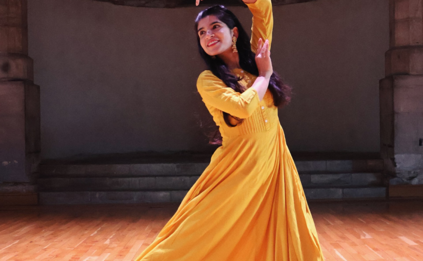 Image of a dancer wearing a long yellow dress moving her arms.