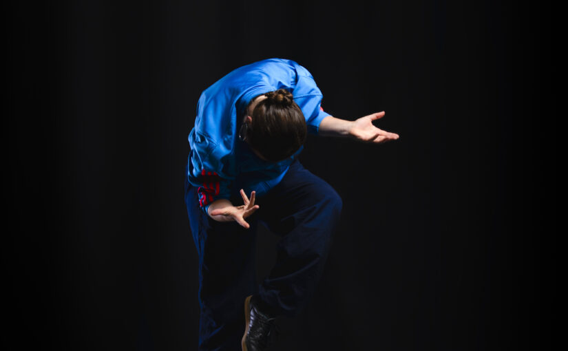 Image of a dancer making a pose wearing a deep blue jacket against a black background.