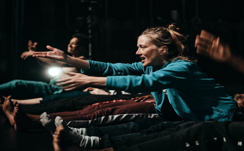 Image of a group od dancers sitting on the floor with their legs and arms extended forward.