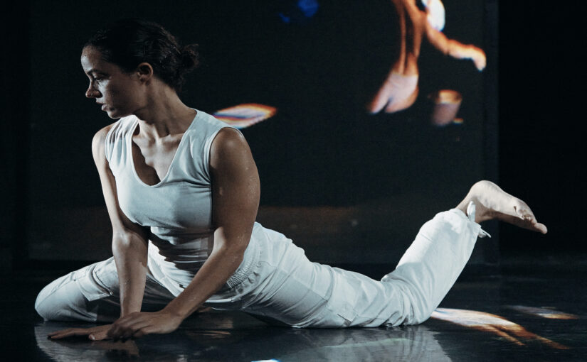 Image of a dancer wearing white clothes on her hands and knees against a dark background.