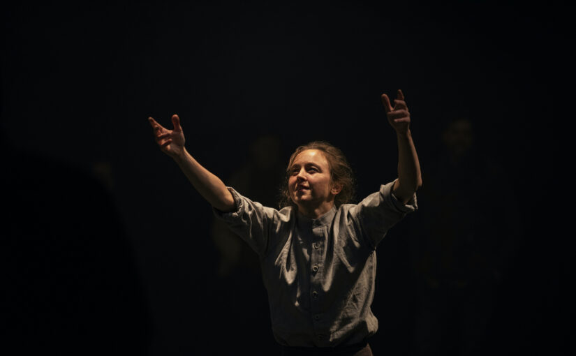 Image of a dancer wearing a light shirt and lifting her arms against a dark background.