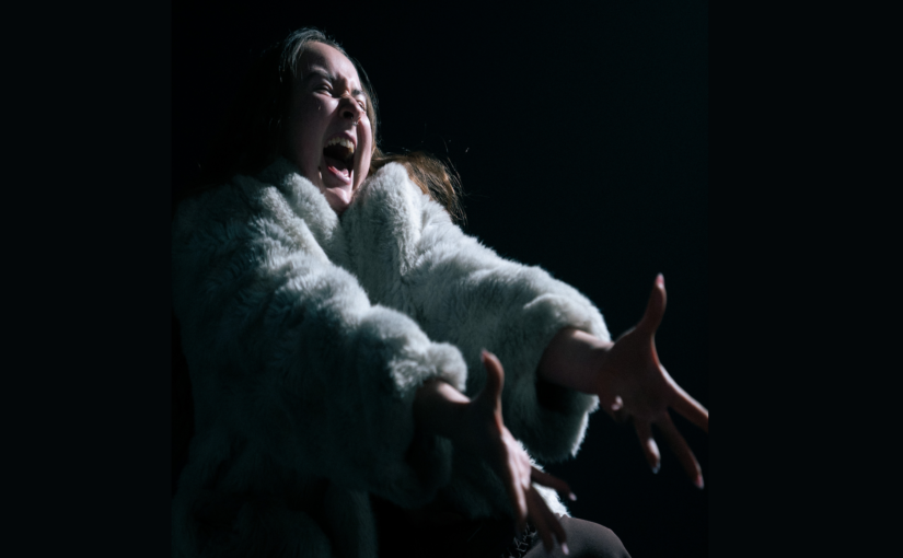 Image of a dancer wearing a coat and extending her arms towards the front while making a shouting expression with her face.