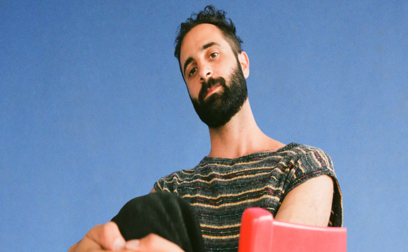 Headshot of a man with a dark beard and short hair, wearing a striped t-shirt against a light blue background.