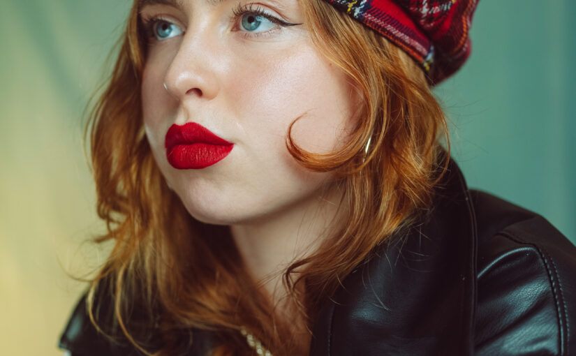 Headshot of a woman with long red hair and red lips wearing a red tartan cap and a black leather jacket.