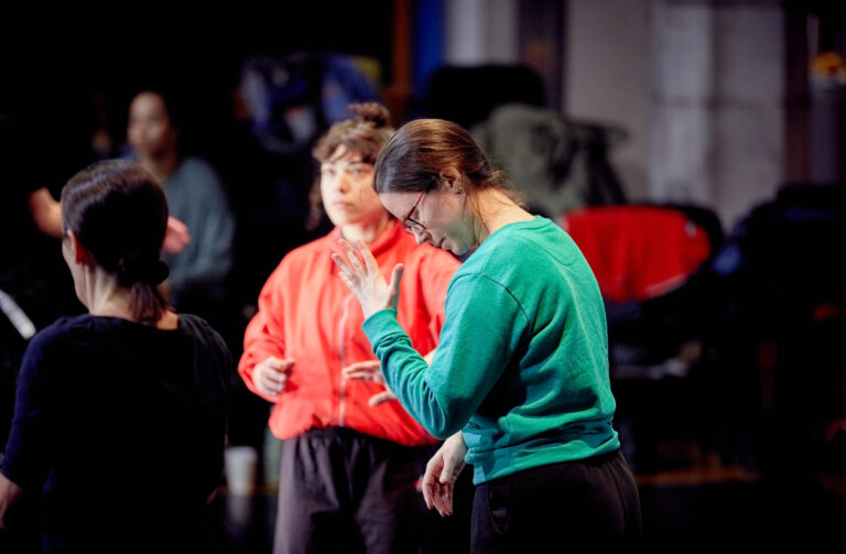 Photo of two dancers in the studio. One is wearing a teal jumper and another one is wearing a coral jacket. The coral coloured letters read percolate 25.