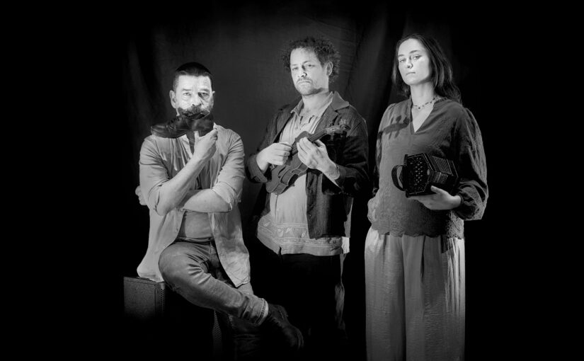 Black and white photo of three performers. One is sitting down holding a dance shoe, the second one is standing up holding a violin and the third one is standing up holding a concertina.